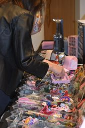 Person in black leather jacket browsing colorful embroidered patches, pins and hair accessories organized in clear bins on a vendor table at a craft market.
