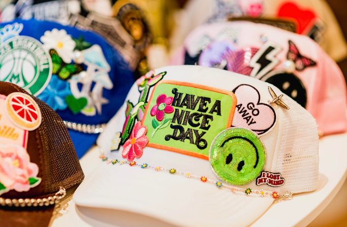 White mesh trucker hat on display covered in colorful embroidered patches — bright "Have a Nice Day" patch, glittery green smiley, "Go Away" heart and floral accents, with other decorated caps blurred in the background.