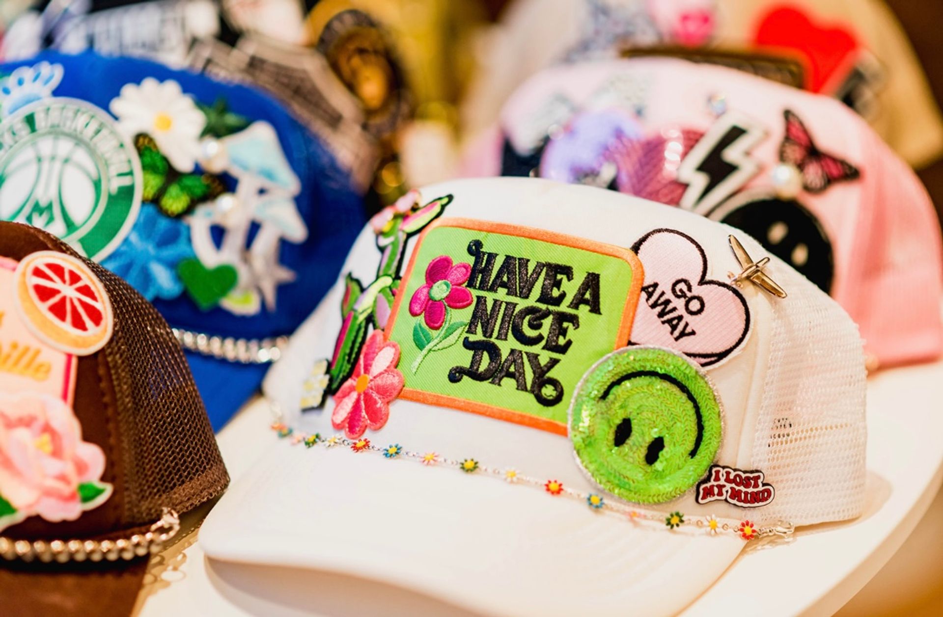 White mesh trucker hat on display covered in colorful embroidered patches — bright "Have a Nice Day" patch, glittery green smiley, "Go Away" heart and floral accents, with other decorated caps blurred in the background.