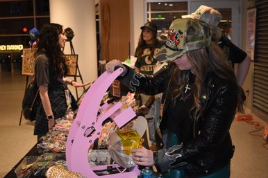 Person using a pink heat-press to customize a mesh trucker hat at an indoor pop-up market vendor table with jewelry and accessories