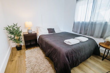 Sunlit modern guest bedroom with queen bed, dark-gray duvet, folded towels, bedside lamp, potted plant and hardwood floor
