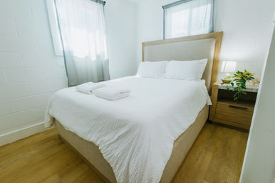Bright, airy bedroom with white linens on a queen bed, wooden headboard and nightstand with lamp and potted plant, light wood floor and sheer curtains