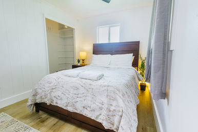 Cozy bright modern guest bedroom with dark wooden headboard, white linens and folded towels on the bed, bedside lamp, potted plant, open closet shelving and gray curtains over hardwood floor