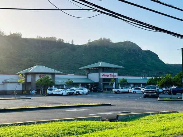 Sunlit green-roofed shopping center with parked cars and a pharmacy storefront in a large lot, framed by a lush green mountain ridge under a clear sky.