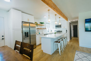 Airy modern farmhouse kitchen with large white island and four metal bar stools, white shaker cabinets, stainless-steel refrigerator, gold pendant lights hanging from an exposed wooden beam, light wood floors and open-concept layout.