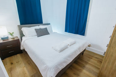 Cozy bright bedroom with crisp white bedding and two folded towels on the bed, gray upholstered headboard, navy curtains, wooden nightstand with lamp and small plant, and light oak floors.