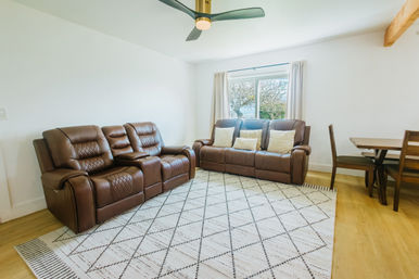 Cozy sunlit living room with brown leather reclining sofas and matching loveseat, cream geometric area rug on light wood floors, small wood dining set, modern ceiling fan, and a window with a tree view.