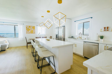 Bright modern coastal kitchen with white island and bar stools, geometric gold pendant lights, stainless appliances and ocean view through large windows.