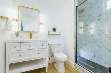 Bright modern white bathroom with gold fixtures: vanity with square mirror, potted plant, toilet, and glass-enclosed marble shower with dark hex tile floor.