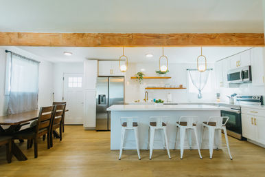 Bright, airy modern farmhouse kitchen and dining area with a large white island and four white metal bar stools, wooden dining table, exposed wood beam, pendant lights, stainless steel appliances, open shelving, and light wood floors.