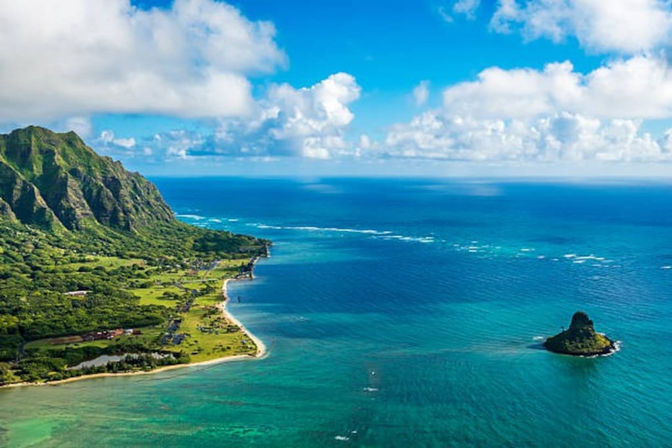 Aerial view of a Hawaiian coastline with turquoise water, sandy beach, lush volcanic cliffs and a small offshore islet under blue skies