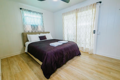 Bright, airy bedroom with a dark purple comforter, white pillows and folded towels on the bed, patterned sheers on a window and sliding glass door, light wood floors and white paneled walls.