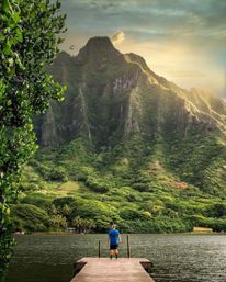 Solo figure on a wooden dock gazing across a calm lake toward towering, lush tropical mountains and golden sunset light