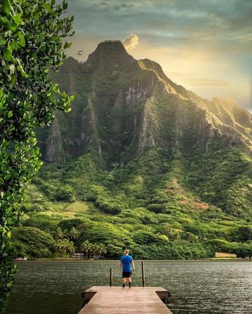 Solo figure on a wooden dock gazing across a calm lake toward towering, lush tropical mountains and golden sunset light