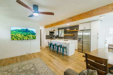 Bright, airy open-concept kitchen and living room with exposed wood beam, island seating with teal bar stools, stainless steel appliances, wall-mounted TV, ceiling fan, and light wood floors.