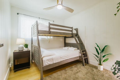 Bright, cozy modern bedroom with a gray wooden bunk bed (double lower, single upper), white linens, angled ladder, wooden nightstand with lamp and plant, ceiling fan, hardwood floor and potted greenery.