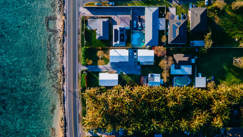 Aerial view of a sunlit coastal neighborhood: turquoise ocean beside a two‑lane beachfront road, houses with pools and solar panels, and a dense palm tree grove.