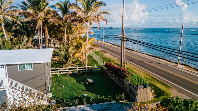 Aerial view of a sunny tropical coastline: palm trees lining an oceanfront two‑lane road with power lines, turquoise sea, and a small seaside house with white roof and fenced yard and picnic table.