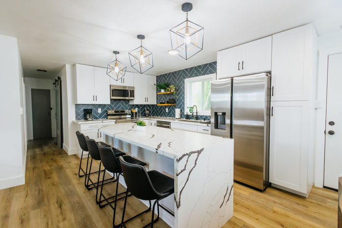 Bright, modern kitchen with a white marble waterfall island, four black bar stools, stainless steel fridge and oven, blue herringbone tile backsplash, geometric pendant lights, white cabinets and warm wood plank flooring.
