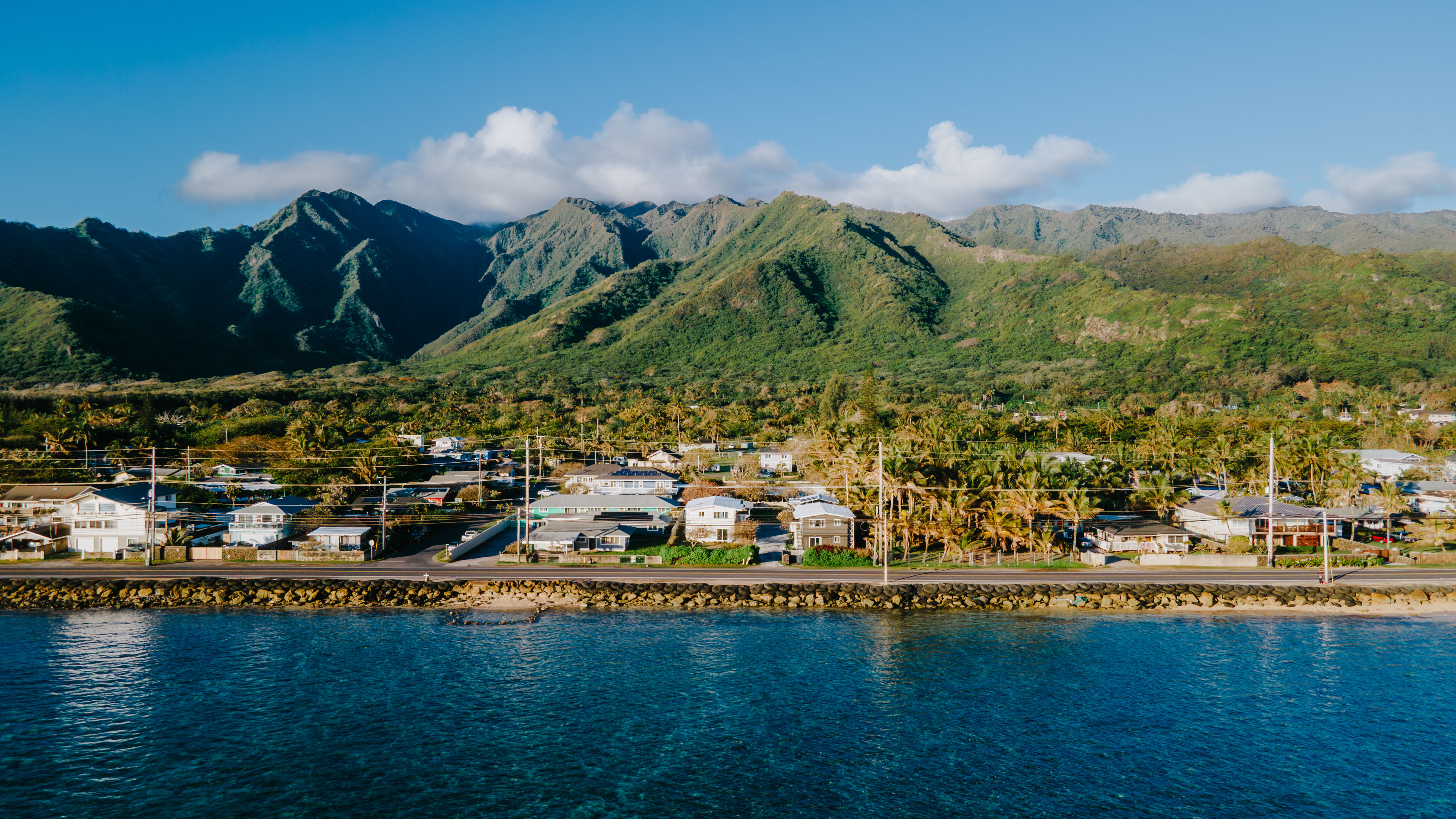 Aerial view of a sunlit tropical coastal town with palm trees and houses along a rocky seawall, deep blue ocean in the foreground and lush green mountains under a bright blue sky.