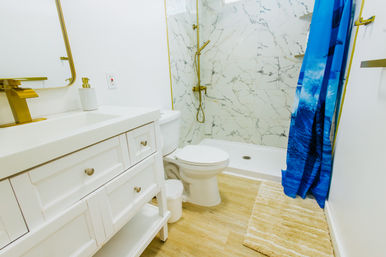 Bright modern bathroom with white vanity and sink, toilet, marble-tiled walk-in shower with gold fixtures, blue ocean-print shower curtain, wood-look floor and beige bath rug.