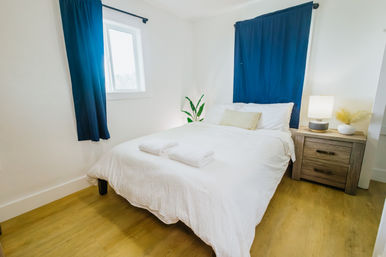 Cozy minimalist bedroom with white linens and folded towels, navy blue curtains, wooden nightstand with lamp and potted plant on warm hardwood floor.