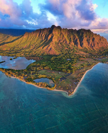 Aerial view of Oahu, Hawaii showing the dramatic Koolau mountain ridgeline glowing at sunset above turquoise lagoon, sandy beach and lush coastal forest