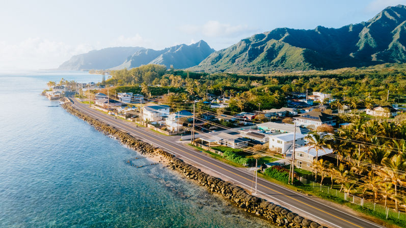 Sunlit aerial view of a tropical coastline: oceanfront highway with rocky seawall, palm-lined beachfront homes and dramatic green mountains in the background