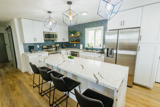 Bright modern kitchen with white cabinets, large veined marble island, four black bar stools, blue herringbone tile backsplash, stainless steel appliances, geometric pendant lights, and light wood floors.