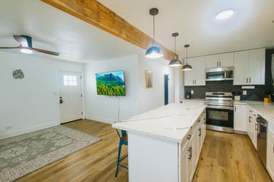 Bright open-plan modern kitchen and living area with a large white marble island, white shaker cabinets, stainless steel oven and microwave, black herringbone tile backsplash, exposed wood beam and three pendant lights, wall-mounted TV by the entry door, blue bar stool and light oak floors.