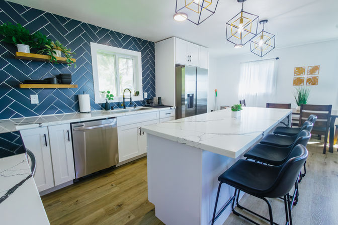 Modern bright kitchen with navy blue herringbone tile backsplash, white shaker cabinets, stainless steel refrigerator and dishwasher, large white marble-look island with four black bar stools, geometric pendant lights, open wood shelves with plants, and light wood flooring.