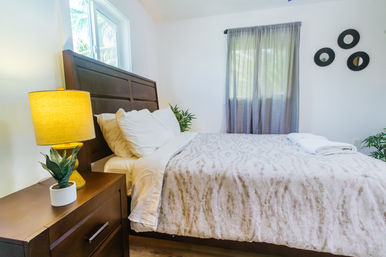 Cozy modern guest bedroom interior with dark wooden headboard, white pillows and neutral patterned bedding, bedside table with yellow lamp and small potted plant, folded white towels on the bed, sheer gray curtains and three round wall mirrors.