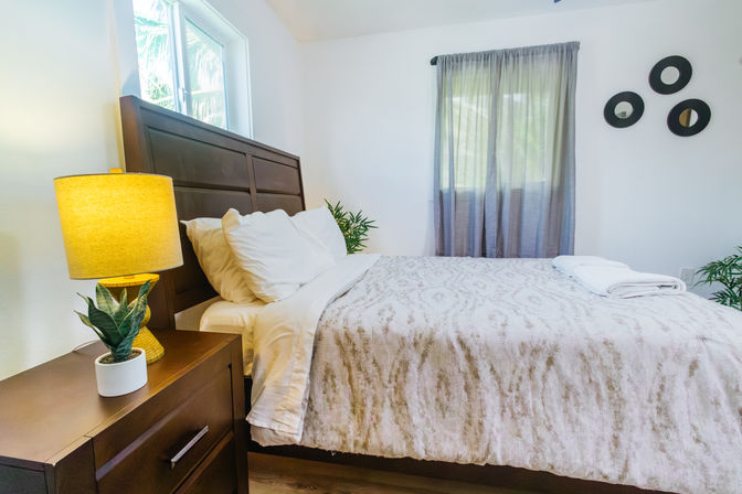 Cozy modern guest bedroom interior with dark wooden headboard, white pillows and neutral patterned bedding, bedside table with yellow lamp and small potted plant, folded white towels on the bed, sheer gray curtains and three round wall mirrors.