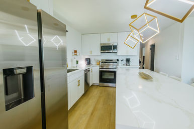Bright modern kitchen with a large white marble island, stainless steel refrigerator and oven, white cabinets with gold hardware, geometric gold pendant lights and light wood floors.