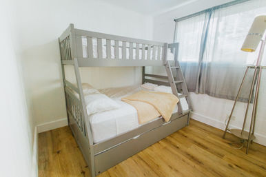 Bright, cozy small bedroom with a gray wooden bunk bed, white linens and a tan throw on oak floors beside a sheer-curtained window and tripod floor lamp.