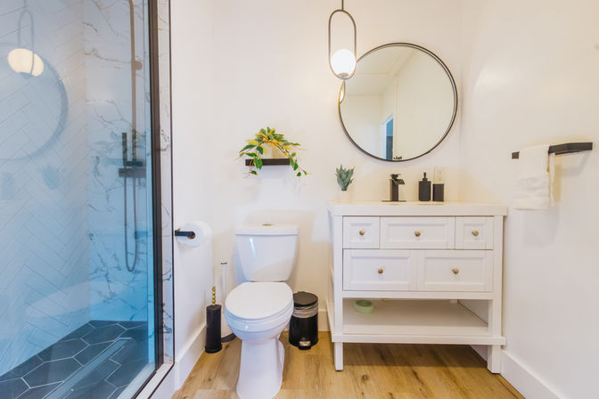 Bright modern white bathroom with glass walk-in shower, black-framed round mirror above a white vanity, white toilet, wood-look floor, black matte fixtures and small potted plants.
