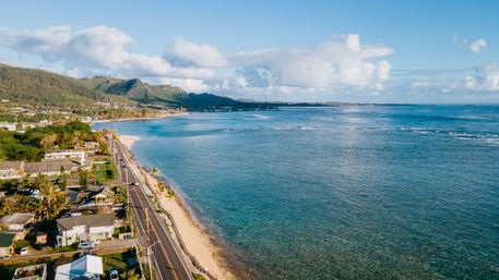 Sunlit tropical coastline with a palm-lined coastal road, sandy beach, clear turquoise ocean and forested green mountains