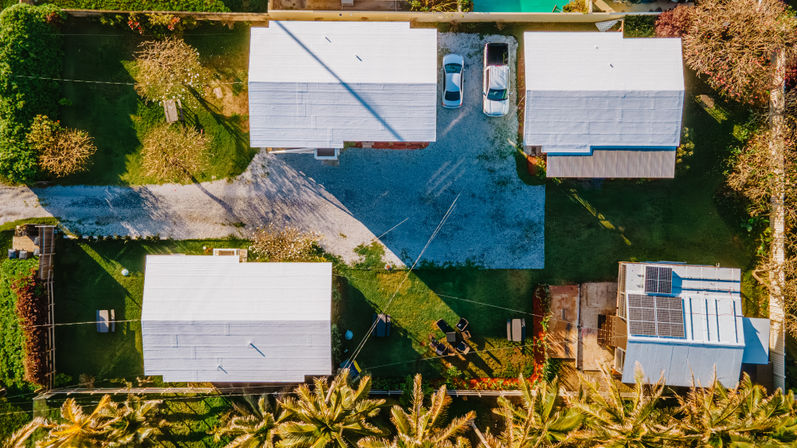 Aerial view of a sunny tropical residential cluster with white-roof bungalows, gravel driveway, green lawns, palm trees, parked cars, and rooftop solar panels.