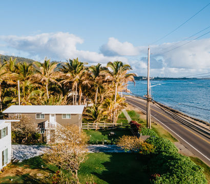 Aerial view of a sunny, palm-lined two-lane coastal highway running beside blue ocean, with beachfront houses, green lawns and distant mountains under fluffy clouds.