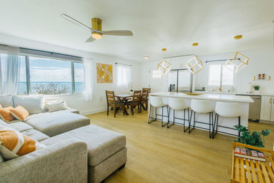 Bright open‑plan coastal living room and kitchen with ocean view through a wide window, gray sectional with orange pillows, white island with four bar stools, wood floors and geometric pendant lights.