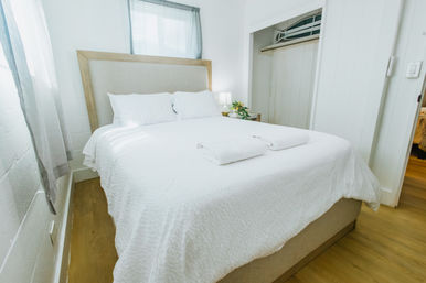 Bright sunlit minimalist bedroom with a neatly made bed in white linens, two pillows and folded towels on top, wooden headboard, bedside lamp and plant, light wood floor and open closet