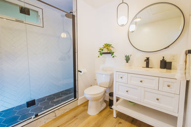 Bright modern white bathroom with glass walk-in shower, round mirror, white vanity, toilet, wood-look floor, and hanging pendant light