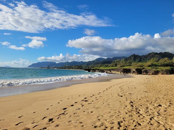 Sunlit tropical beach with sandy shoreline and footprints, turquoise waves lapping rocky coastal cliffs and lush green mountains under a bright blue sky with scattered clouds.