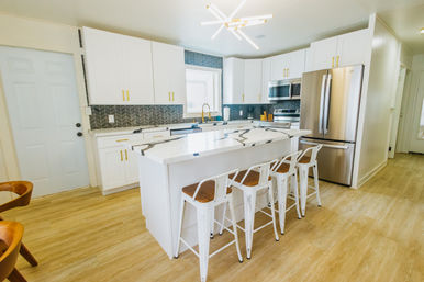 Bright modern kitchen with white cabinets and gold hardware, dark chevron tile backsplash, large white marble island with four white metal bar stools, stainless steel refrigerator and light wood floors.