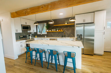 Bright modern kitchen with a marble island, four teal metal bar stools, stainless steel fridge and appliances, dark herringbone tile backsplash, floating wood shelves, pendant lights, exposed wooden beam, and light hardwood floors.