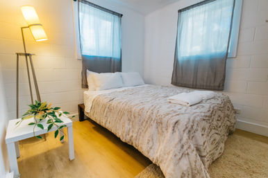 Cozy minimalist bedroom with a double bed in a soft patterned beige comforter, neatly folded towels and white pillows, sheer gray curtains over two windows, warm floor lamp, small white side table with a leafy plant, and light wood floors.