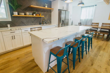 Bright modern kitchen with a large white marble island and four teal metal bar stools, white shaker cabinets, dark herringbone tile backsplash, open wood shelves, stainless refrigerator, and warm wood flooring.