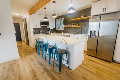 Bright modern open-concept kitchen with large white marble island, four teal metal bar stools, stainless steel appliances, dark herringbone tile backsplash, floating wood shelves, exposed beam and pendant lights on light wood floors