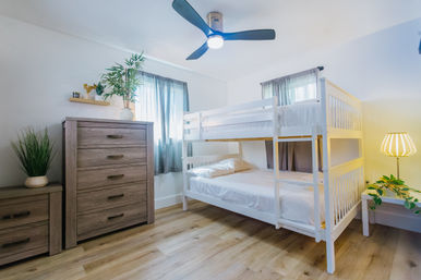 Bright family-friendly bedroom with white wooden bunk bed, light wood dresser and nightstand, ceiling fan, potted plants, sheer curtains and warm bedside lamp on hardwood floors.