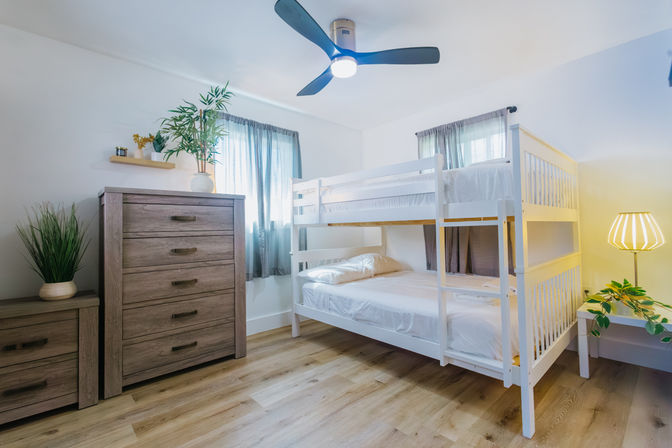 Bright family-friendly bedroom with white wooden bunk bed, light wood dresser and nightstand, ceiling fan, potted plants, sheer curtains and warm bedside lamp on hardwood floors.